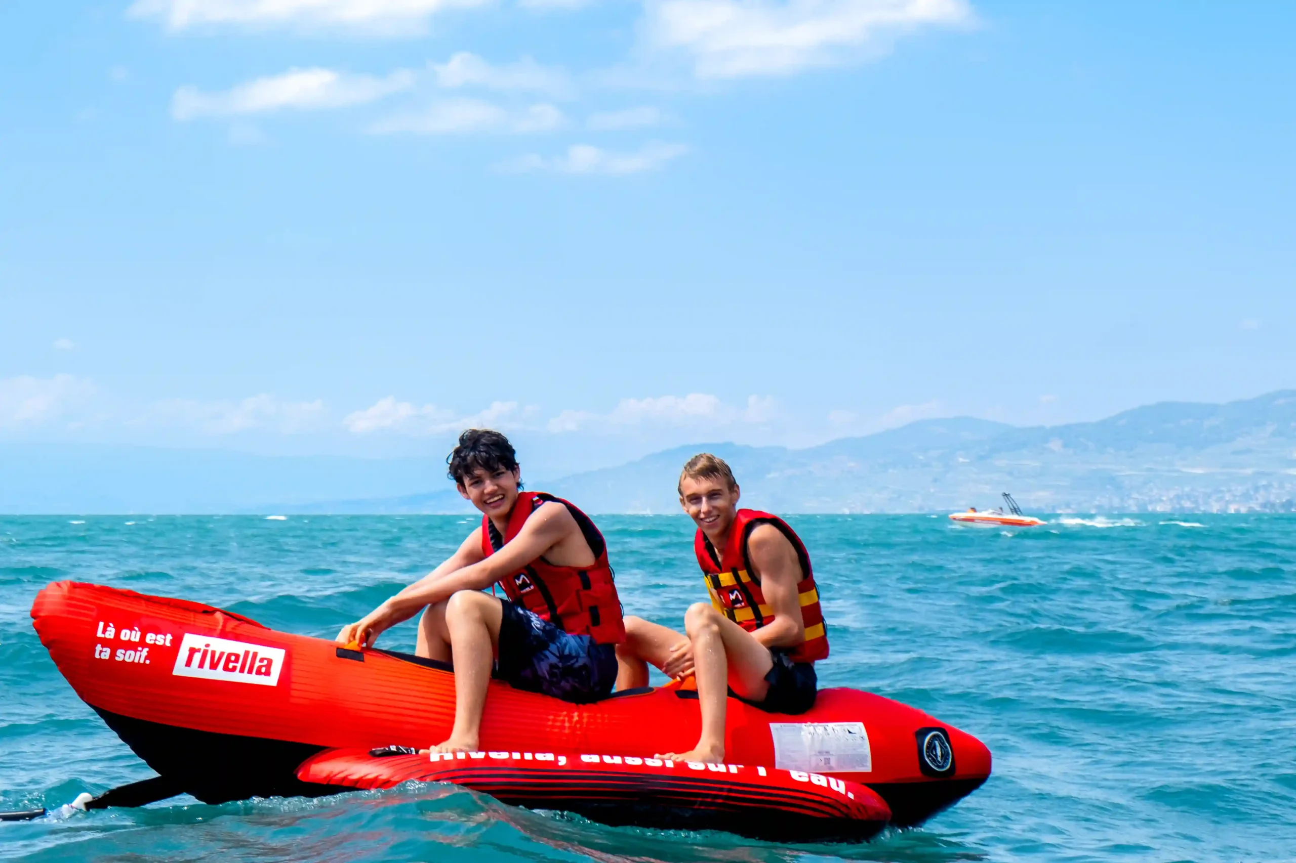 Two teens on a red inflatable boat on a sunny lake, wearing life jackets, mountains in the background.
