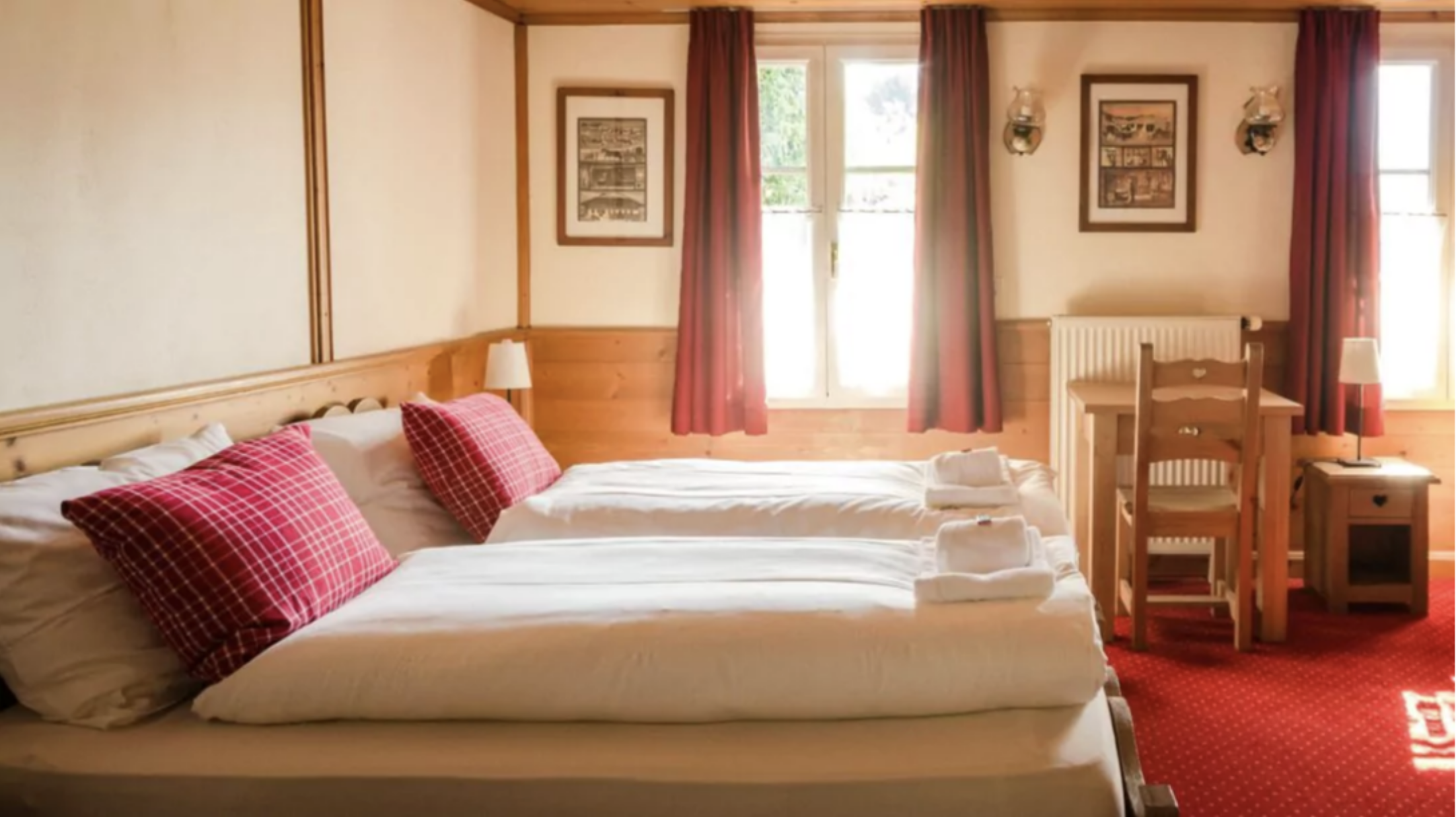 Sunlit alpine hotel room with twin beds, red plaid pillows, wooden furniture and red curtains