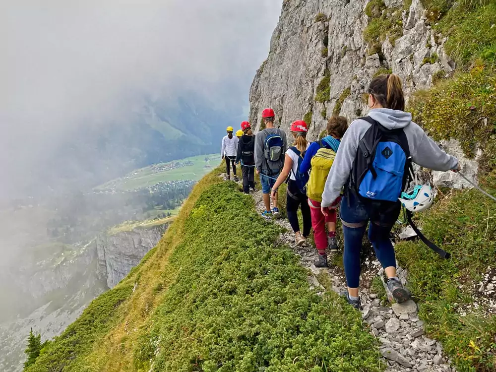 Group of hikers on a narrow cliffside mountain trail with helmets, overlooking a foggy alpine valley