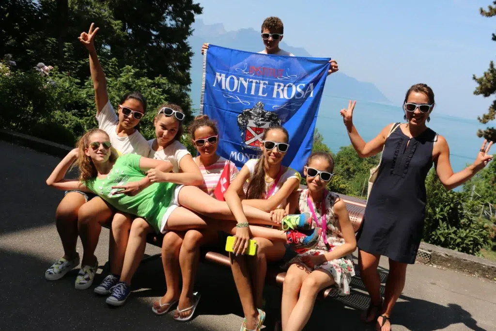 Group of students in sunglasses posing with Institut Monte Rosa banner by a lake and mountains.