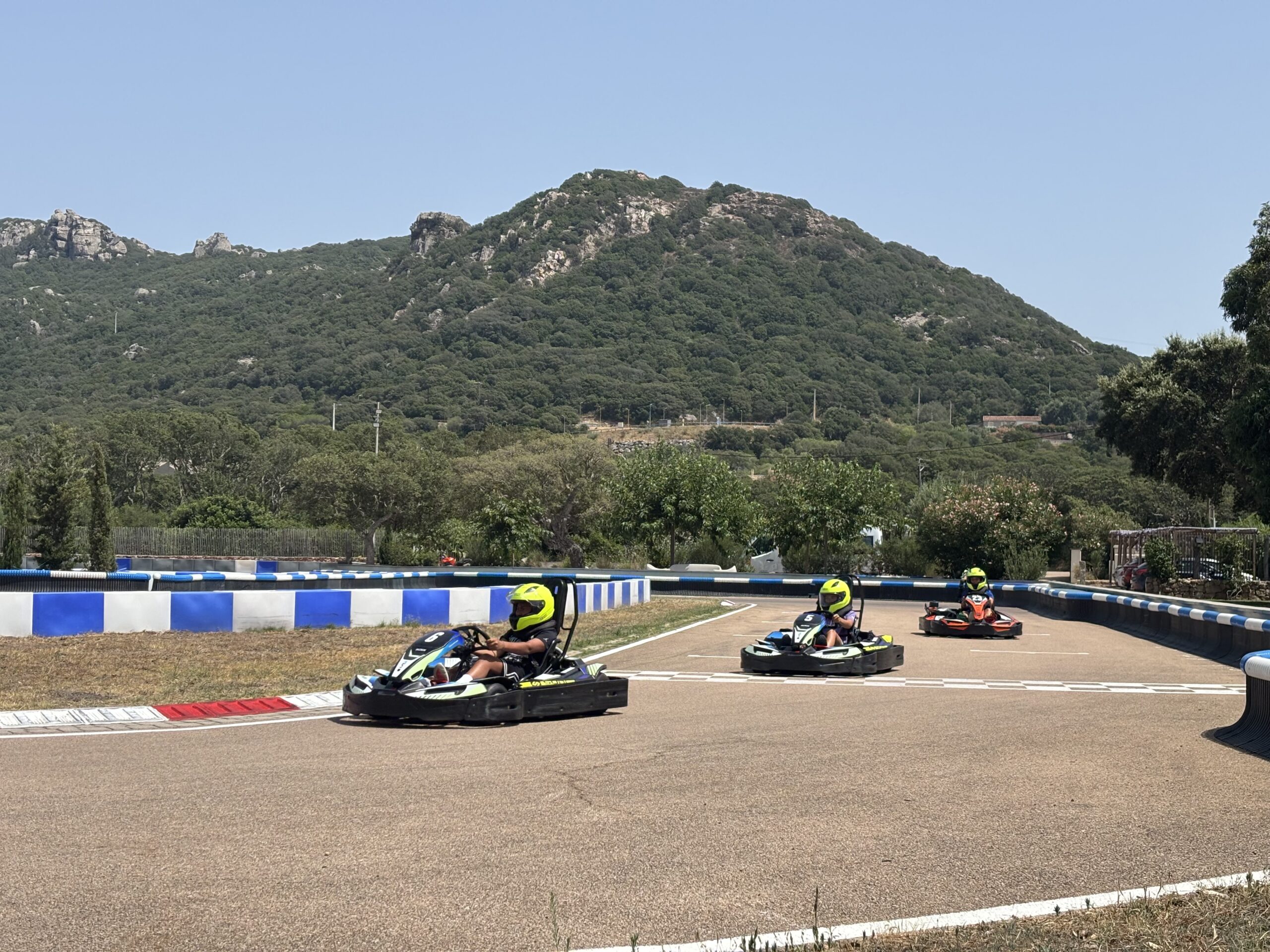 Three go-kart drivers cornering on an outdoor track with blue-white barriers and mountain backdrop
