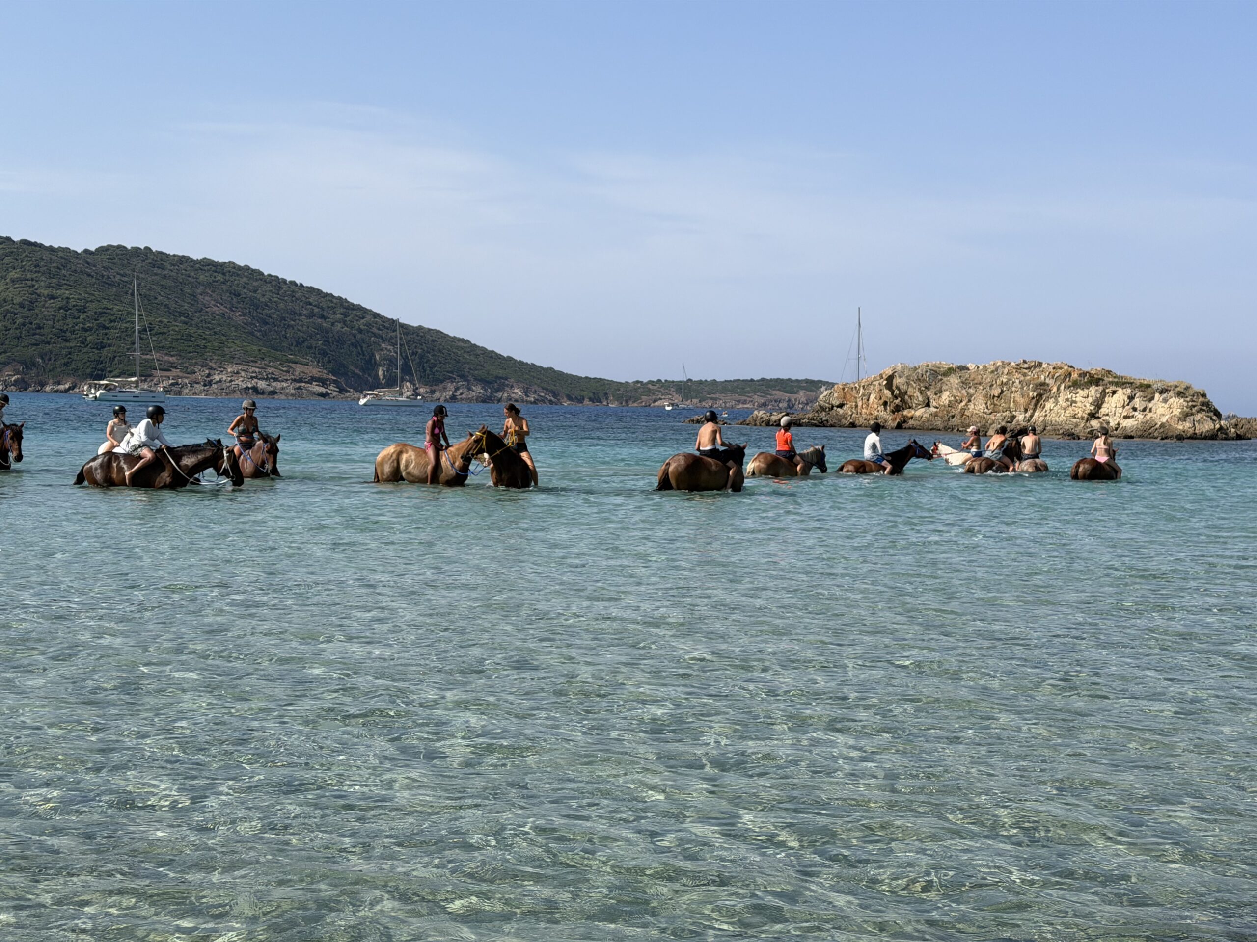 Group riding horses through shallow turquoise water at a Mediterranean beach with sailboats and rocky islet.