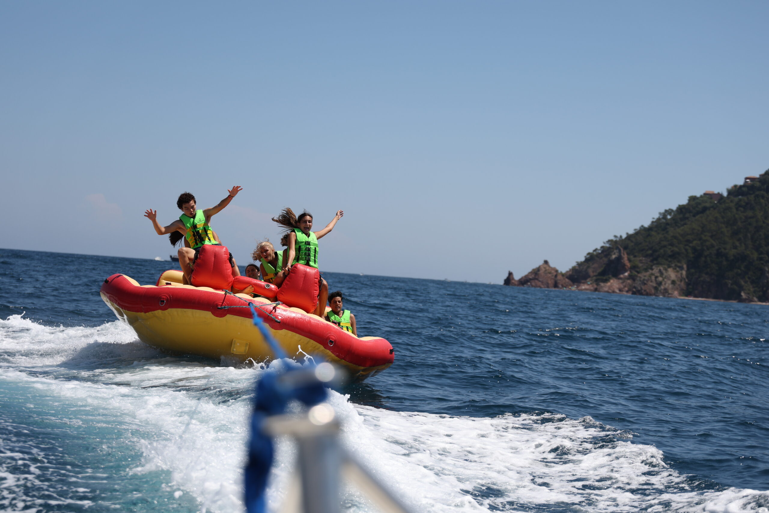 Group riding a towable banana boat on blue sea, wearing life jackets near rocky coastline on a sunny day