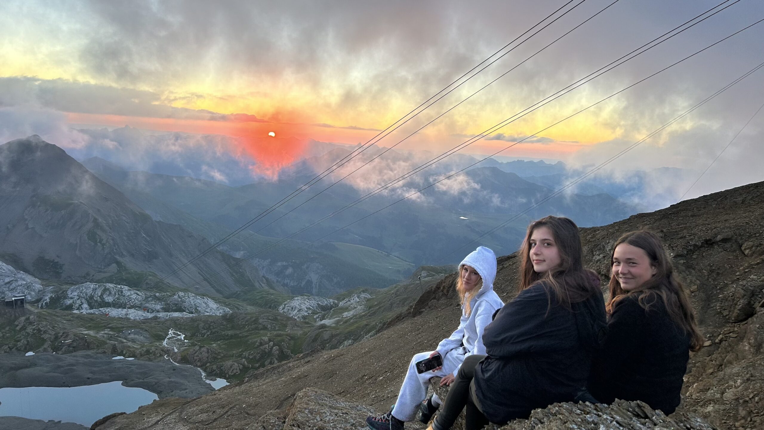 Three hikers on a rocky alpine ridge watch a glowing sunset over misty mountains and cable car lines